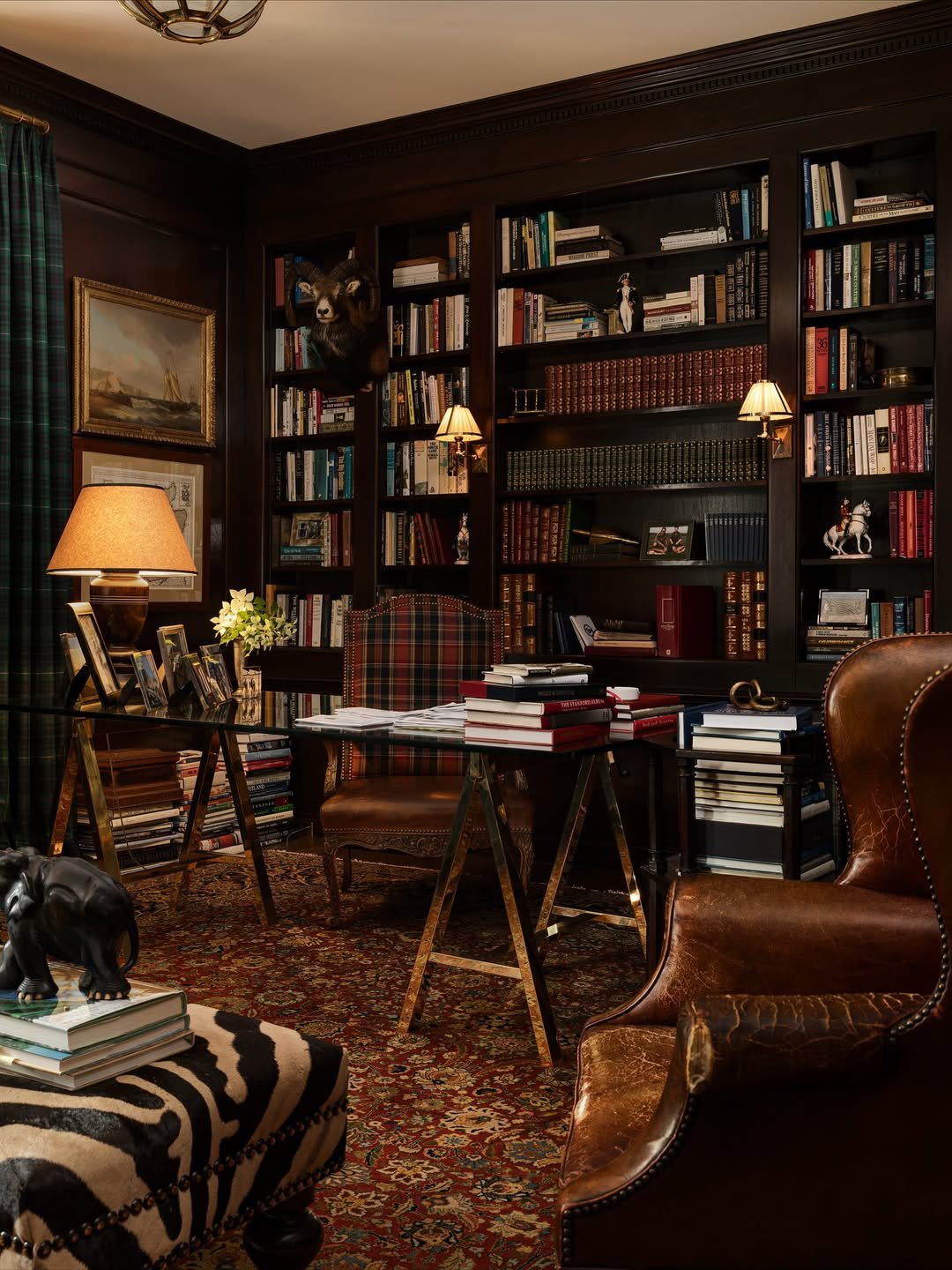 A book-lined study with a writing desk, brass lamp and a hide rug